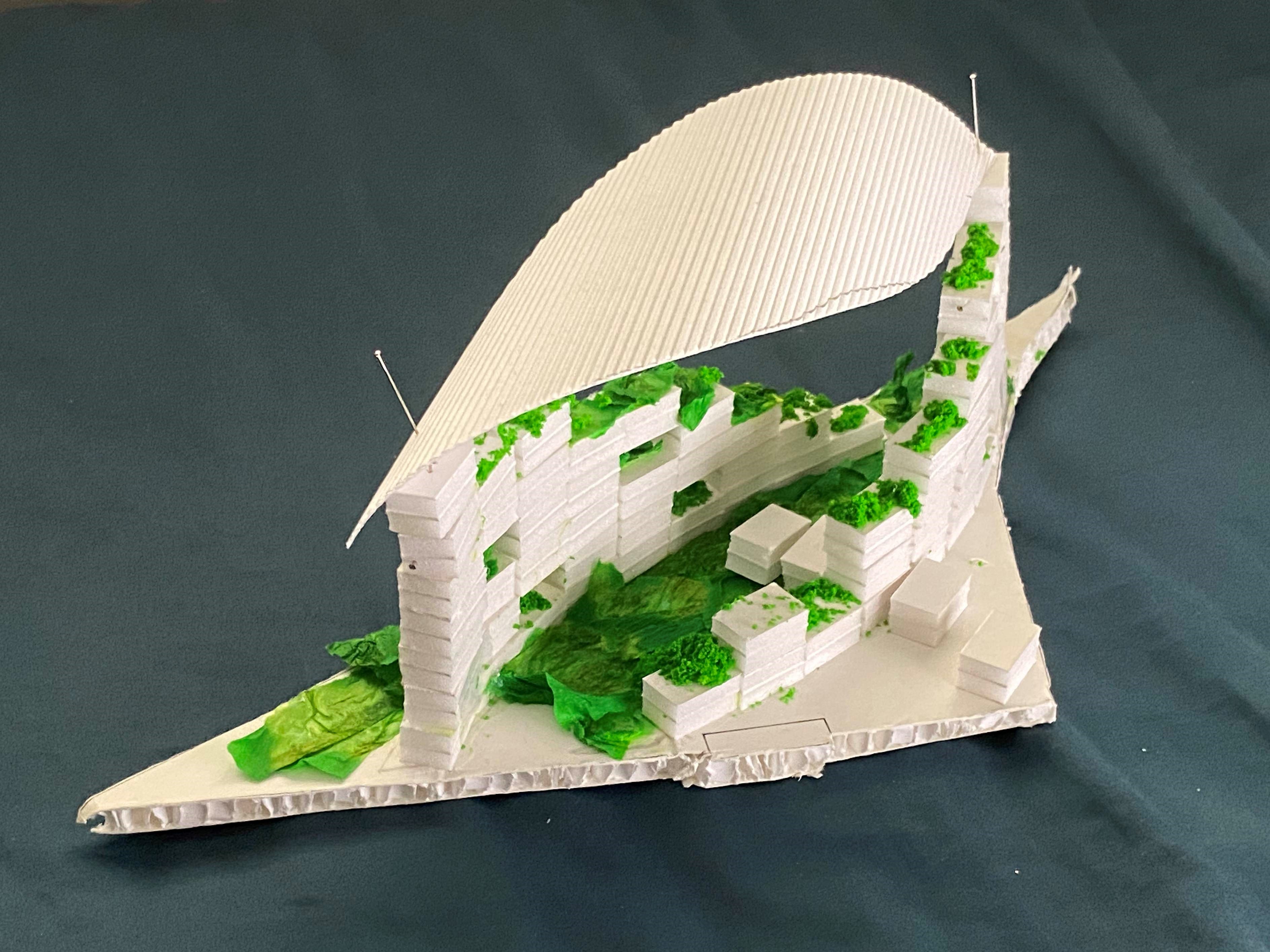 White cardboard architectural scale model with a curved roof, tiered walls, and green foliage accents on a gray surface.